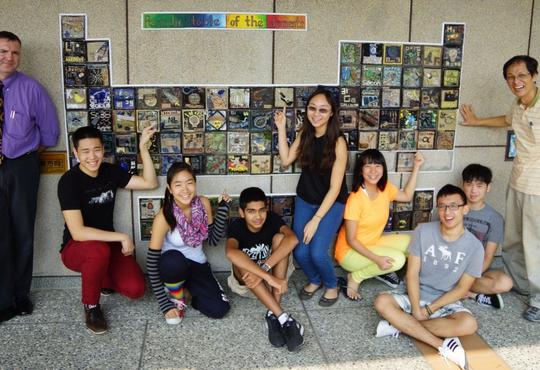 Students and two teachers standing in front of periodic table on wall.