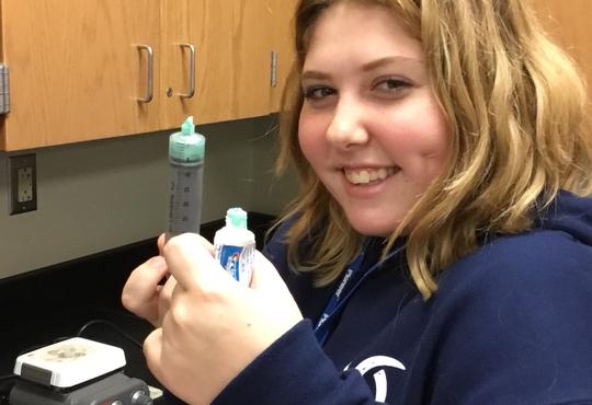A smiling girl squeezing a tube of toothpaste in one hand and the other hand has a syringe.