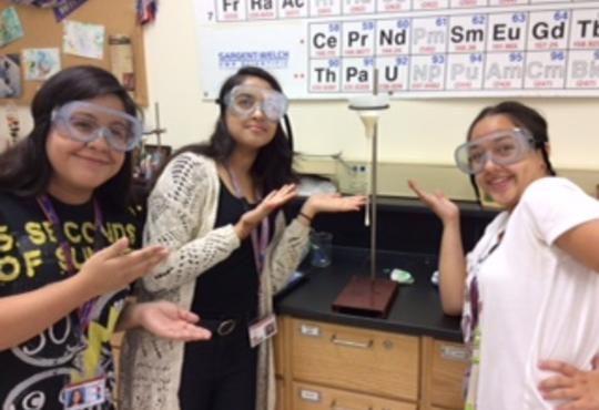 Three girls in a lab pointing to a drop of white material coming out of a funnel