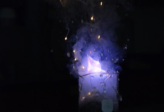 A beaker with a clear and colourless solution with a bright purple flame on top and golden sparks flying off in a darken room