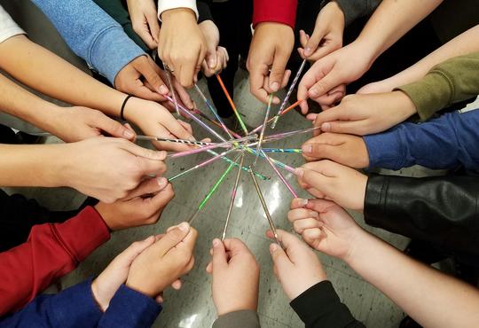 About 20 hands holding stirring rods in a circle. The stirring rods are brightly coloured.