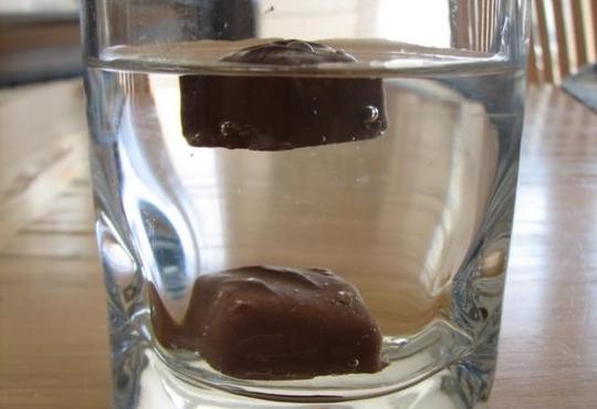 Clear glass of water with two squares of chocolate, one floating, the other one has sunk to the bottom.