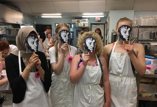 four students in white lab aprons in a chemistry lab holding up black and white Avogadro masks