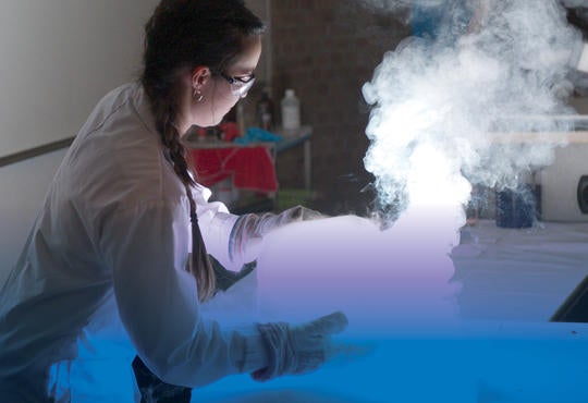  A girl with a lab coat, goggles and large gloves is picking up a white glowing cube with what appears to be white smoke coming 