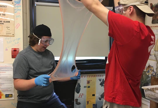two boys with goggles and blue gloves stretching out a piece of slime in the lab