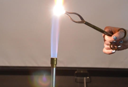 a lit Bunsen burner with hand holding a tongs into the flame resulting in a very bright white light  