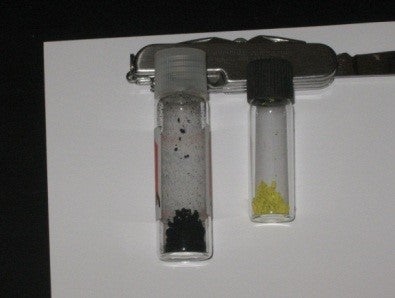 Small vial containing black substance beside small flask containing yellow substance.