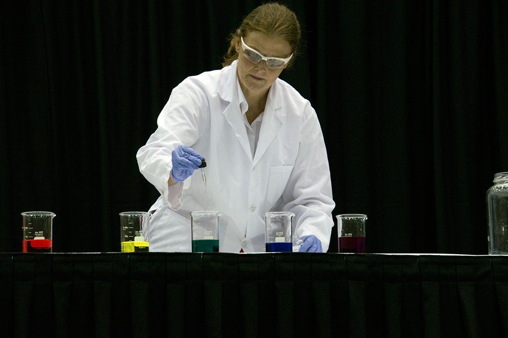 Woman giving chemistry demonstration dripping dyes into beakers.
