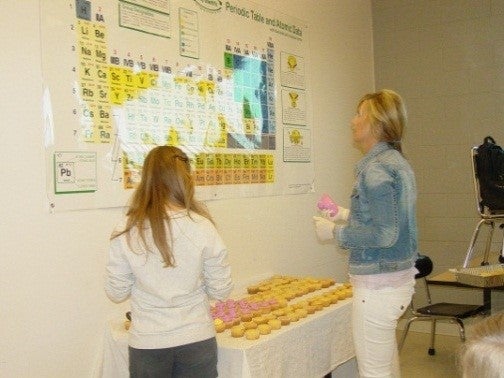 Two females looking at the periodic table and arranging cupcakes, each with a different element symbol in icing, to form a periodic table.