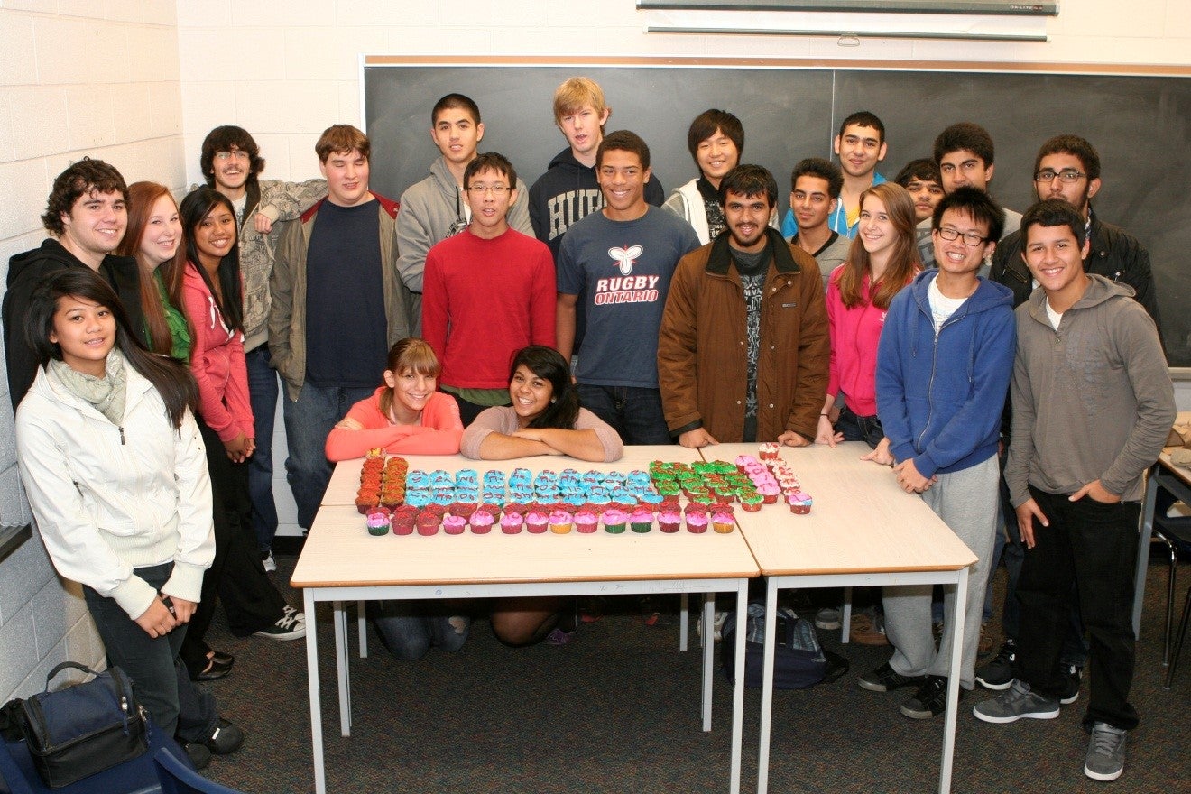 A classroom room of smiling high school students with a periodic table made of cupcakes, with each cupcake having an elemental symbol in icing.