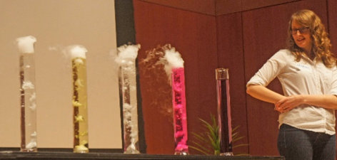 A woman standing next to a table with four out of five graduated cylinders fuming.