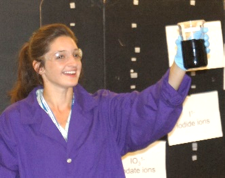 A woman shows a beaker with dark blue/black solution.