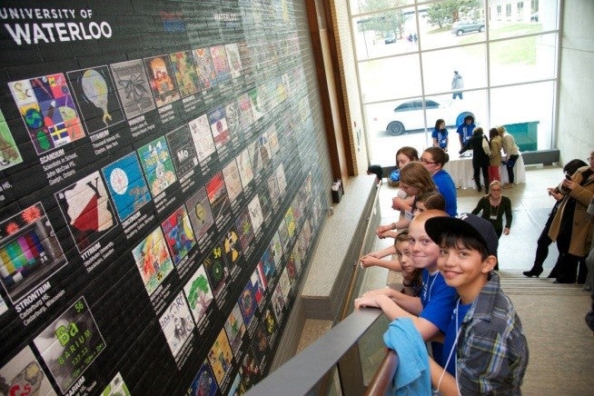 Young teenagers smiling while standing on stairs and looking at the 2-story high Periodic Table Project Wall mural.