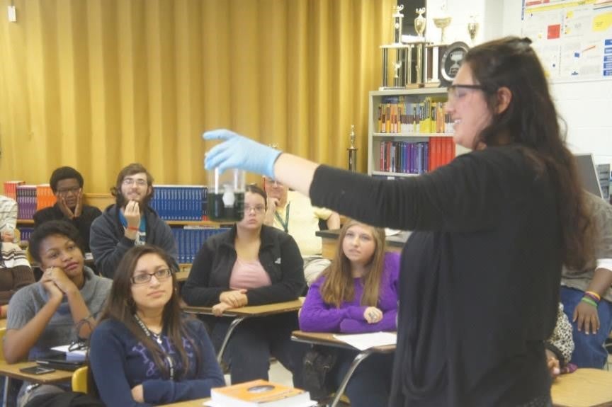 Teacher holding beaker with dark solution in front of classroom.