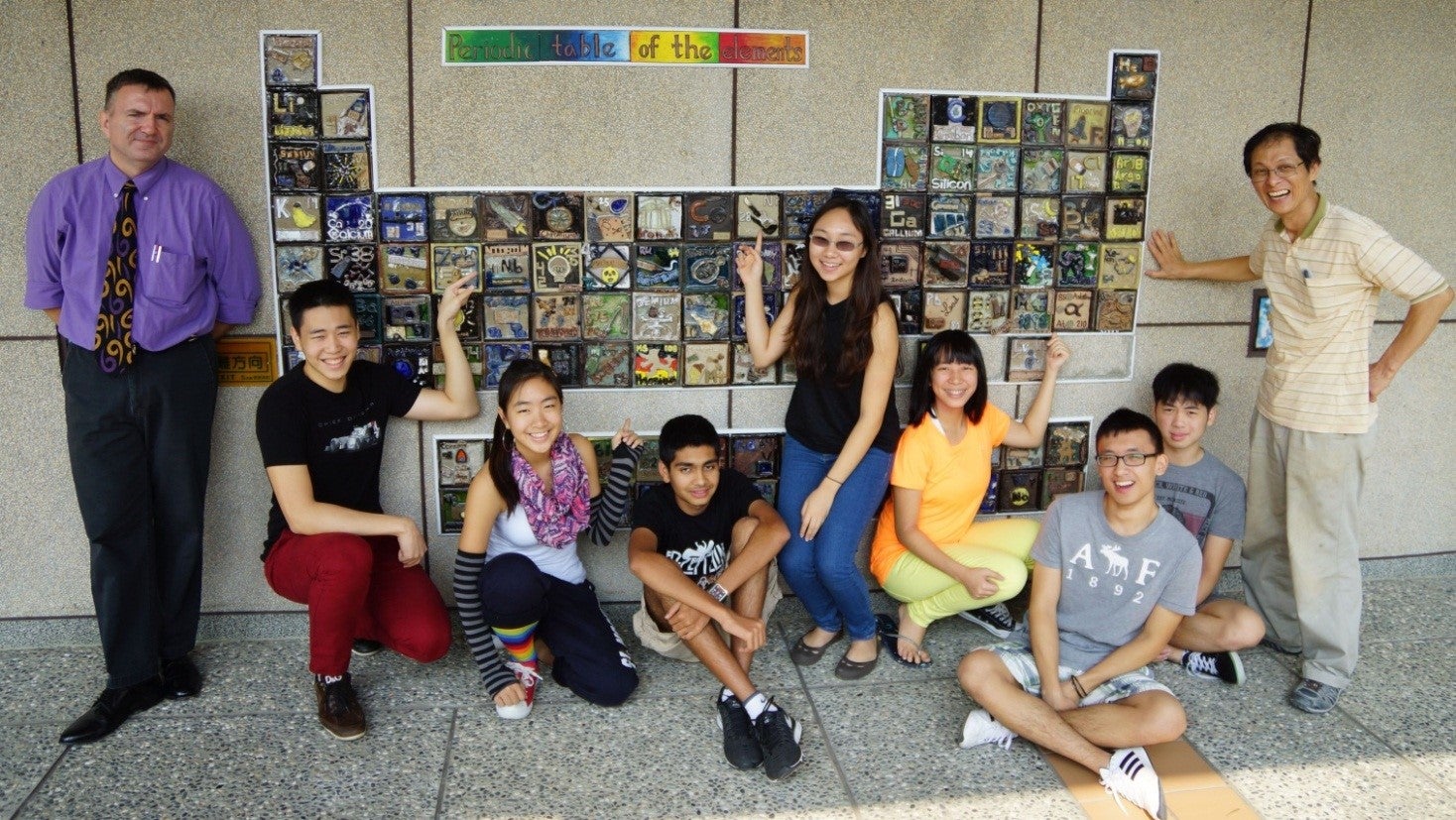 Students and two teachers standing in front of periodic table on wall.