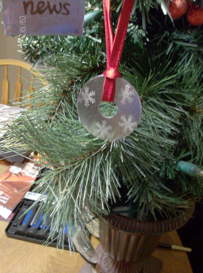 Metal disk etched with snowflakes on ribbon, Christmas tree in background.