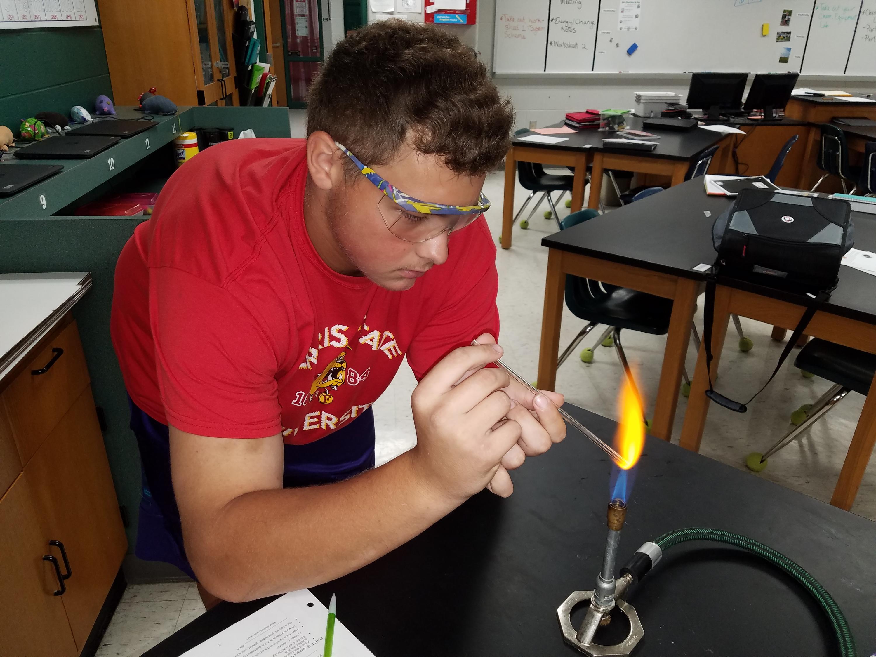 A boy holding a glass rod into the tip of the blue cone of the flame of a Bunsen burner at a 45 degree angle - the flame is bright yellow