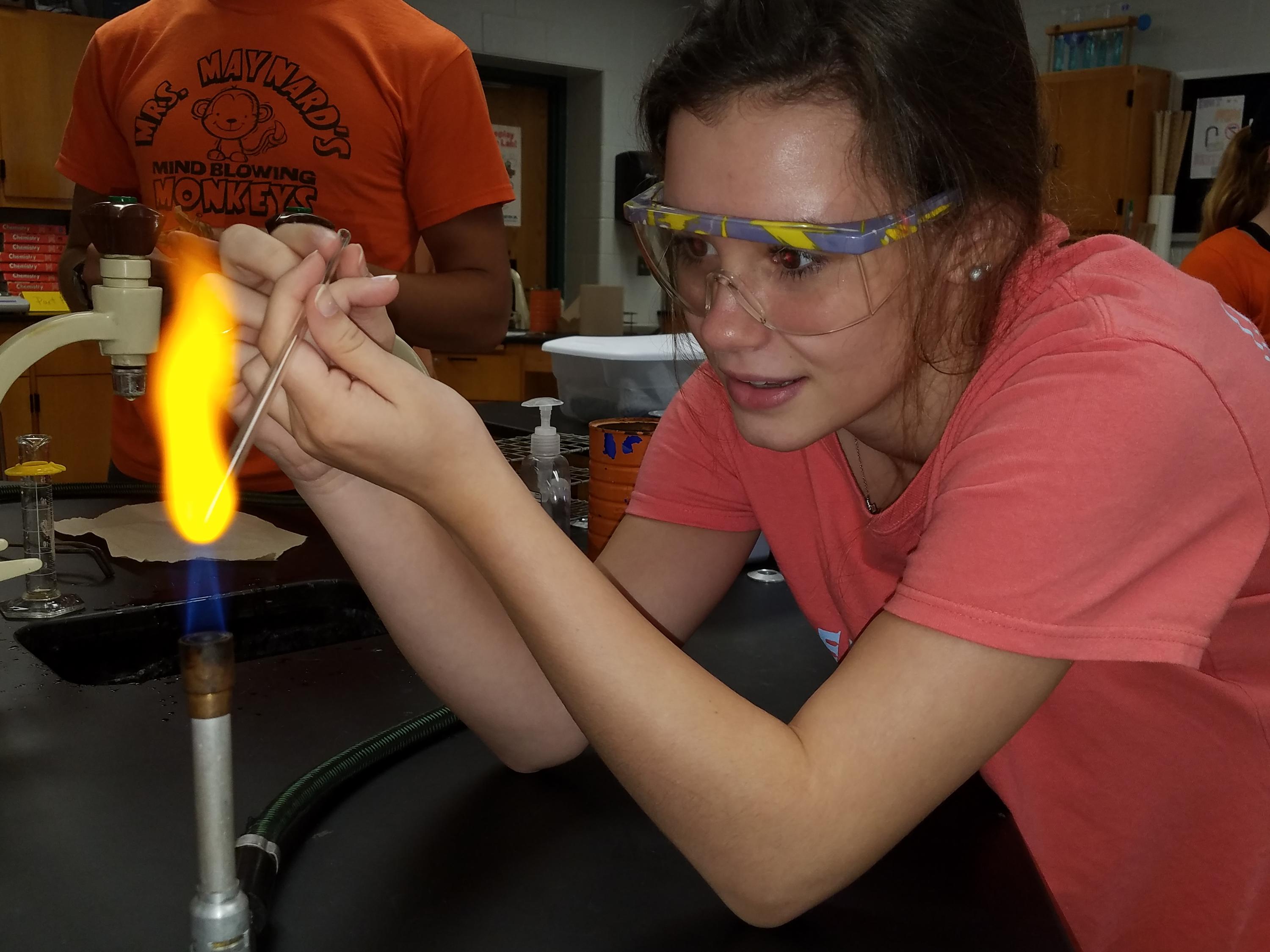 A girl holding a glass rod into the tip of the blue cone of the flame of a Bunsen burmer at at 45 degree angle - the flame is bright yellow