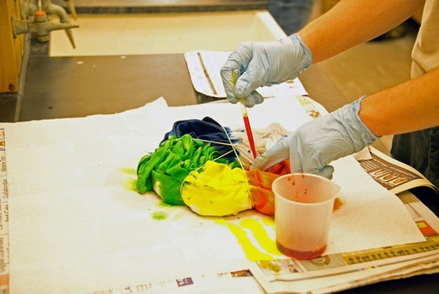 A T-shirt bundled up with elastics and adding orange dye with a pipette. Yellow, green and blue have already been added to the T-shirt.