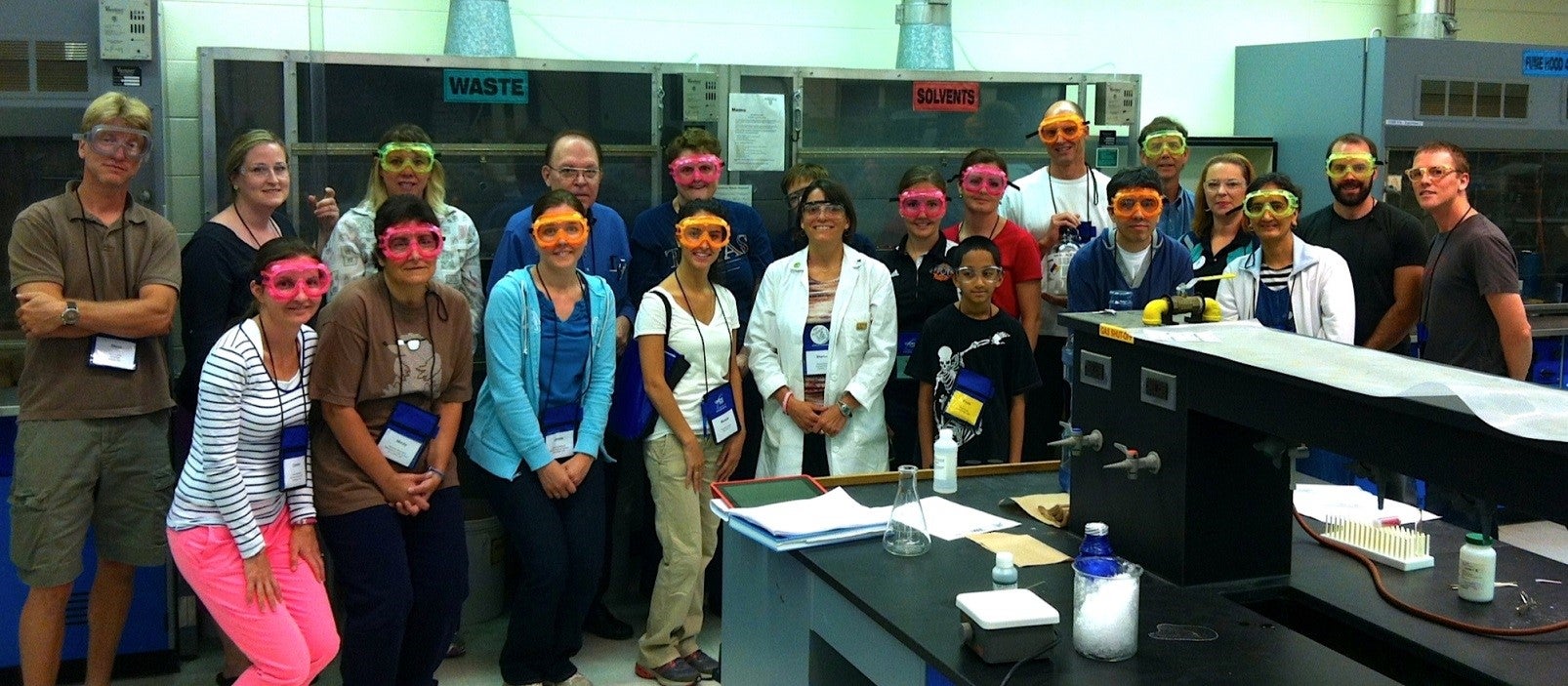 A large group of ChemEd attendees with brightly coloured goggles in front of a fume hood in a lab setting. (Sharon Geyer's session, with Sharon in the front row in a white lab coat).
