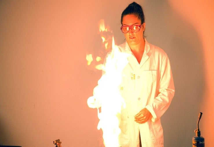 Faye Twiddy in a white lab coat with goggles behind a large flame.