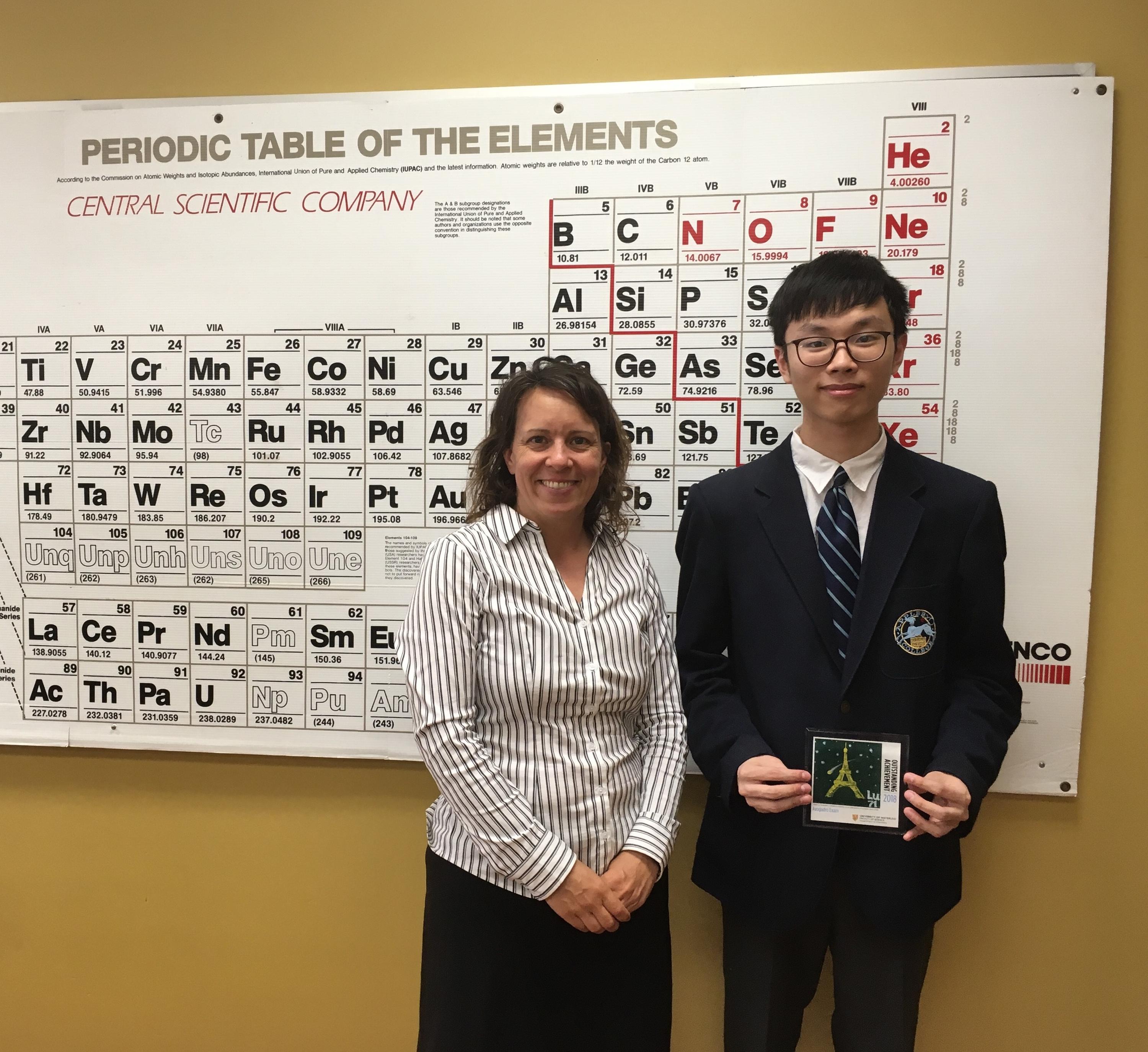 Stephen Chen holding a winning tile in front of a periodic table with teacher, Melissa Rathier