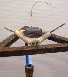 Two bottle cap put together – one is the bottom, the other acts as a cover and a wire holding them together The bottle caps are shown on a clay triangle over a blue Bunsen burner flame.