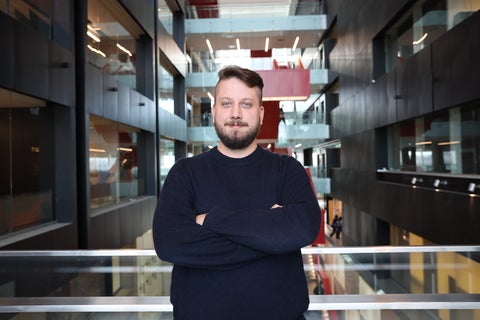 a man with a beard standing with arms crossed in a blue sweater