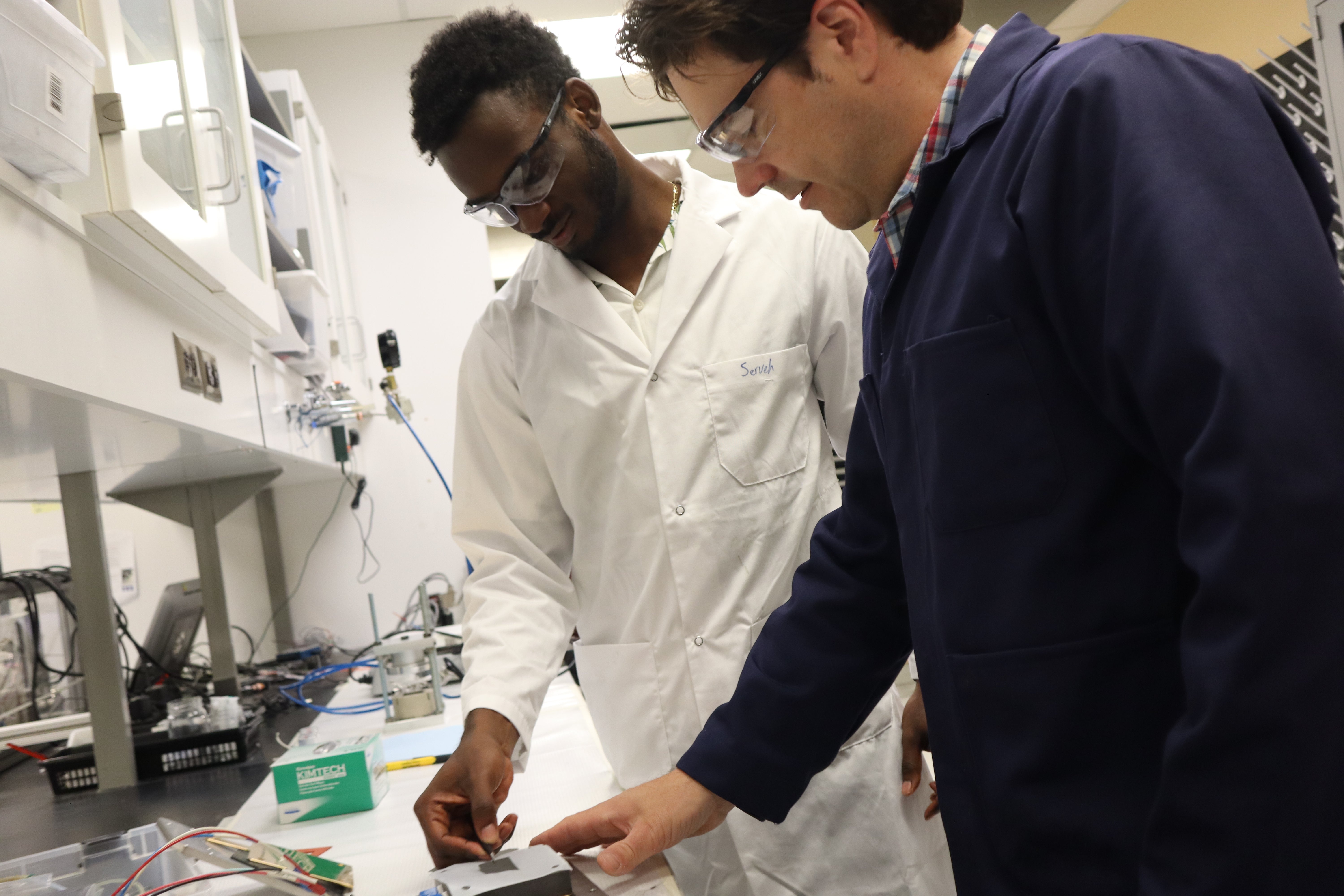 two men in lab coats and safety glasses looking at a battery