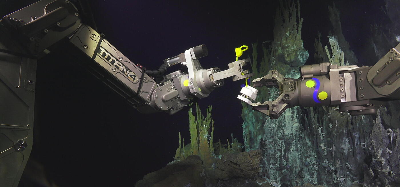 Remote Operating Vehicle arms holding a cube shaped sampling instrument. Background has blue hudrothermal vents covered in small