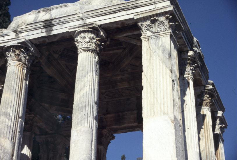 A close of of the corner of the ruins of a Roman temple, focusing on tall, fluted columns with a leaf motive at the top. The ceiling of hte temple is visible through the columns, and is instricately carved.