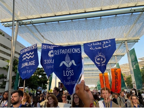Protest signs at COP 28 reading Reparations for damage to water, for stolen land, for loss of livelihood
