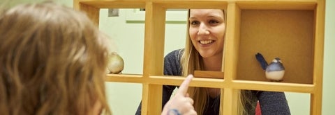 A clinical psychology peers through a wooden bookshelf at a young child as they engage in play activity