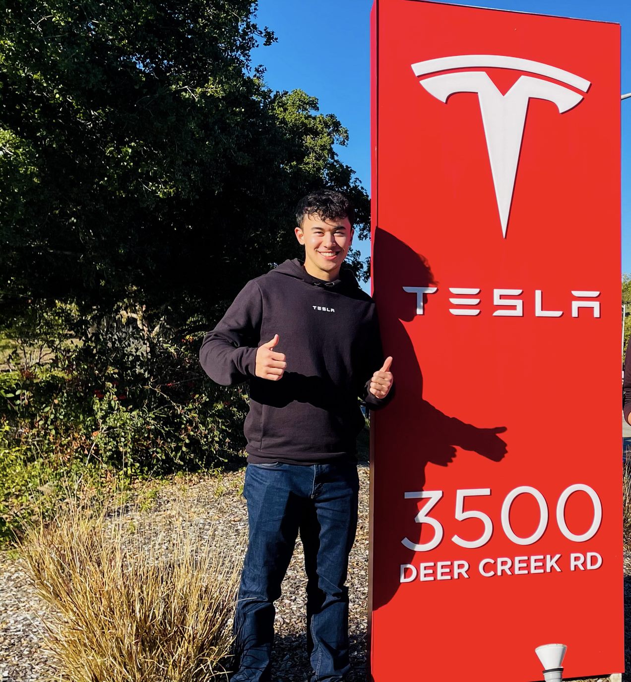 A photo of Brandon smiling and giving two thumbs up beside a Tesla sign.