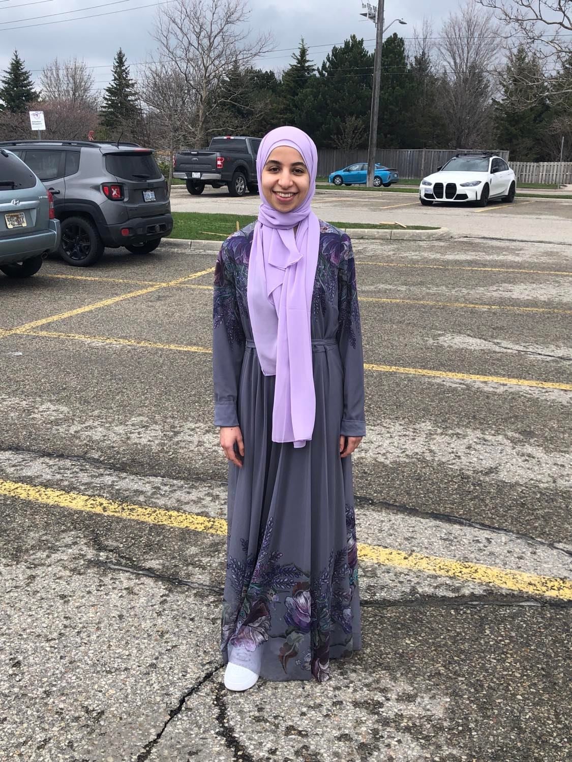 A photo of Fatima standing and smiling in a parking lot wearing a light purple hijab.