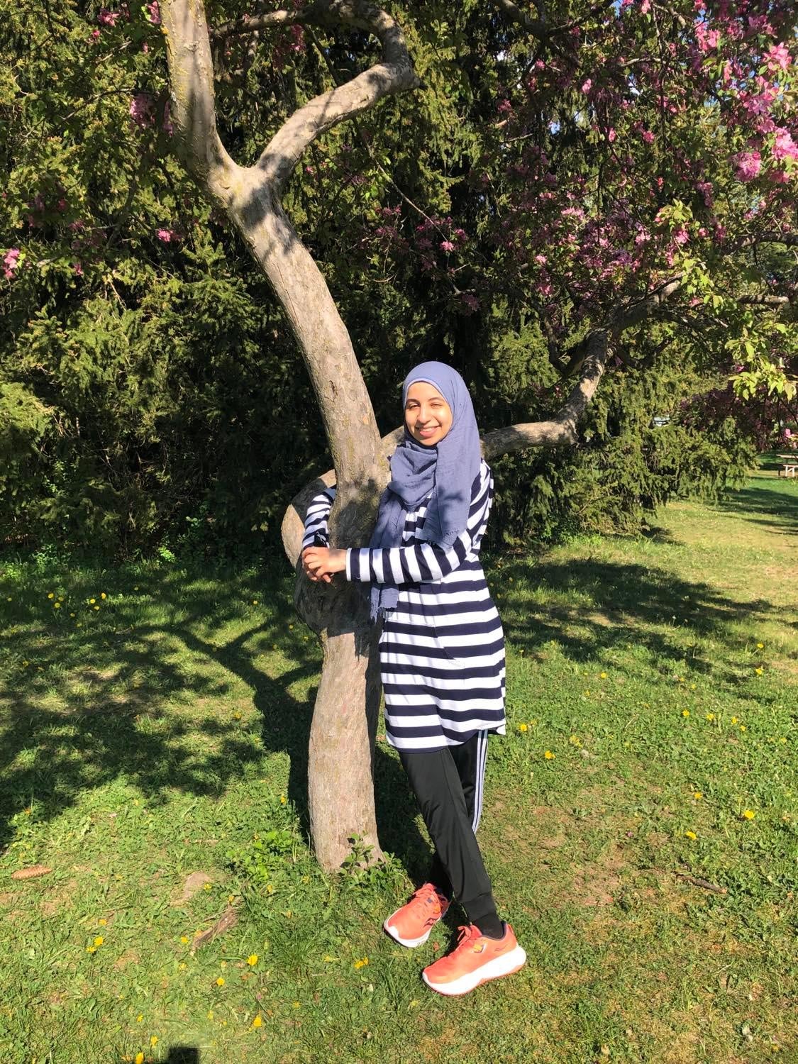 A photo of Fatima standing and smiling with her arms wrapped around a tree.