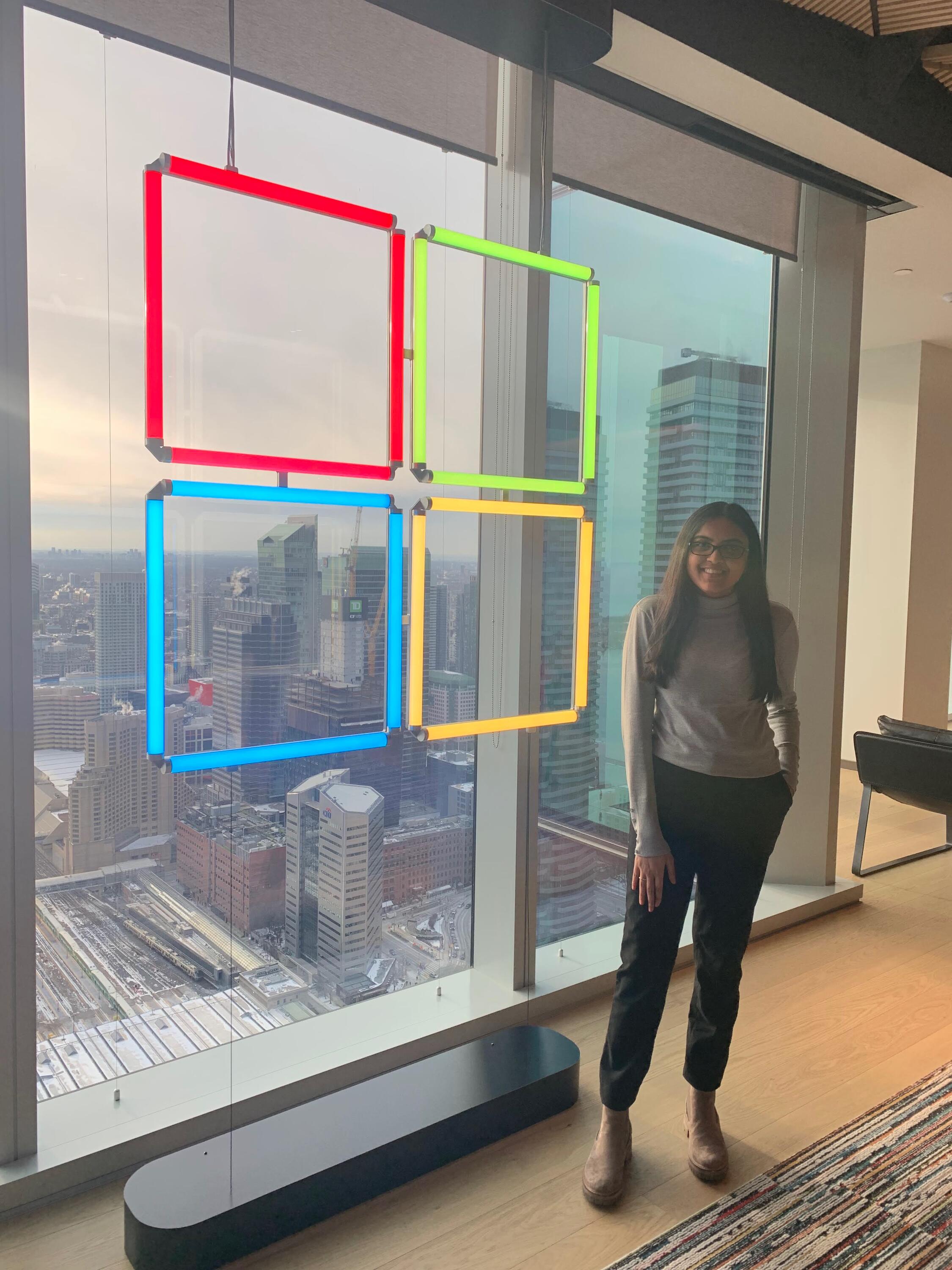Dhruvi standing beside a window with the Microsoft logo handing infront