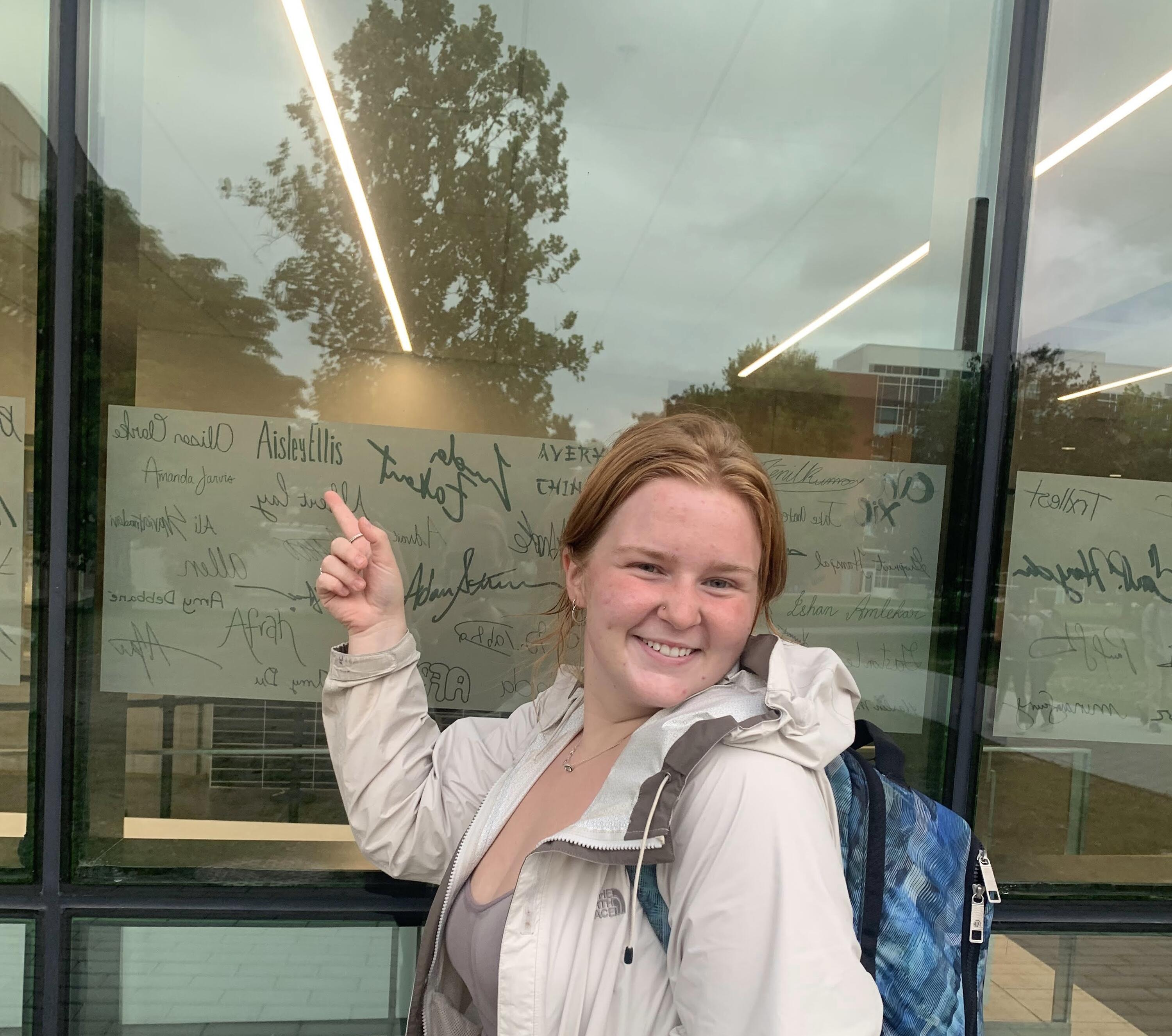 Aisley Ellis pointing to her written name on the window of Student Life Centre.