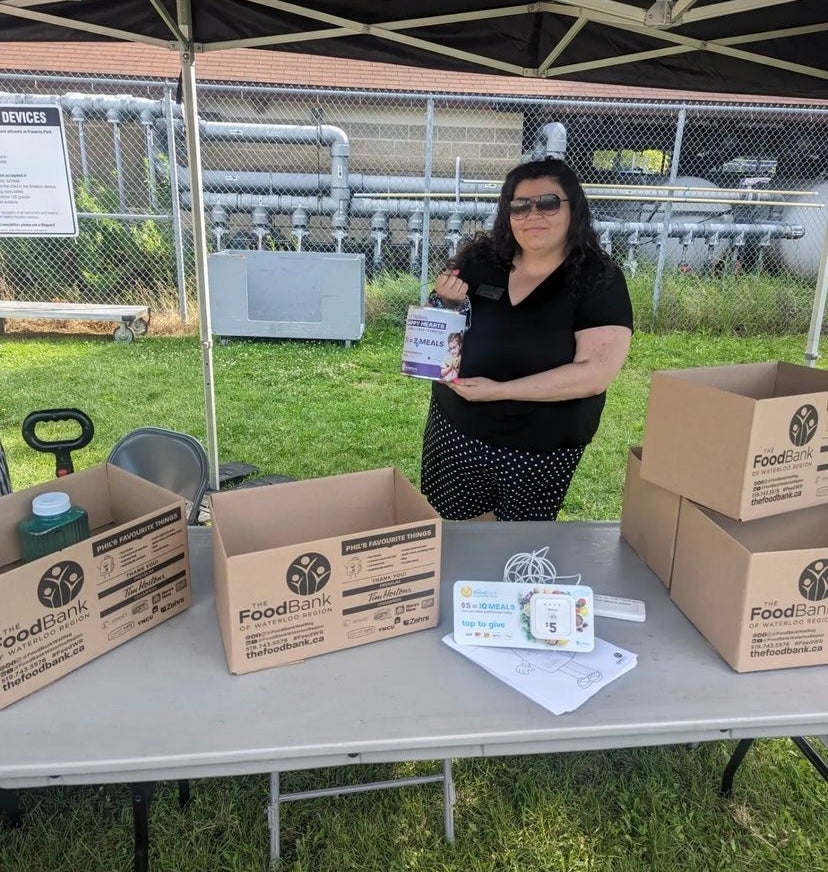 Vanessa Cerqueira standing under a tent, behind a table with cardboard boxes, holding a donations can.
