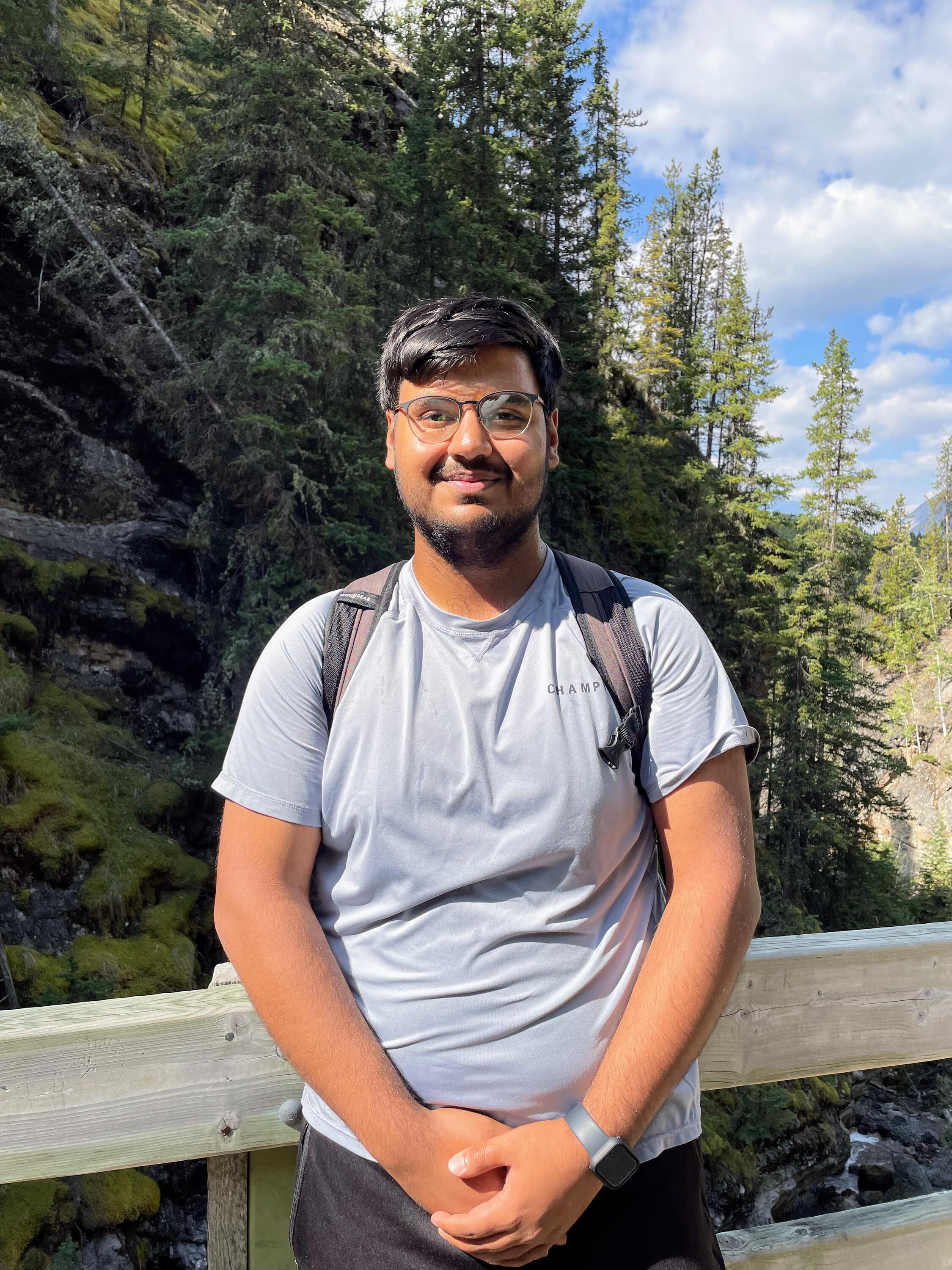 Shashwat smiling in a forest and leaning against a wooden railing. 