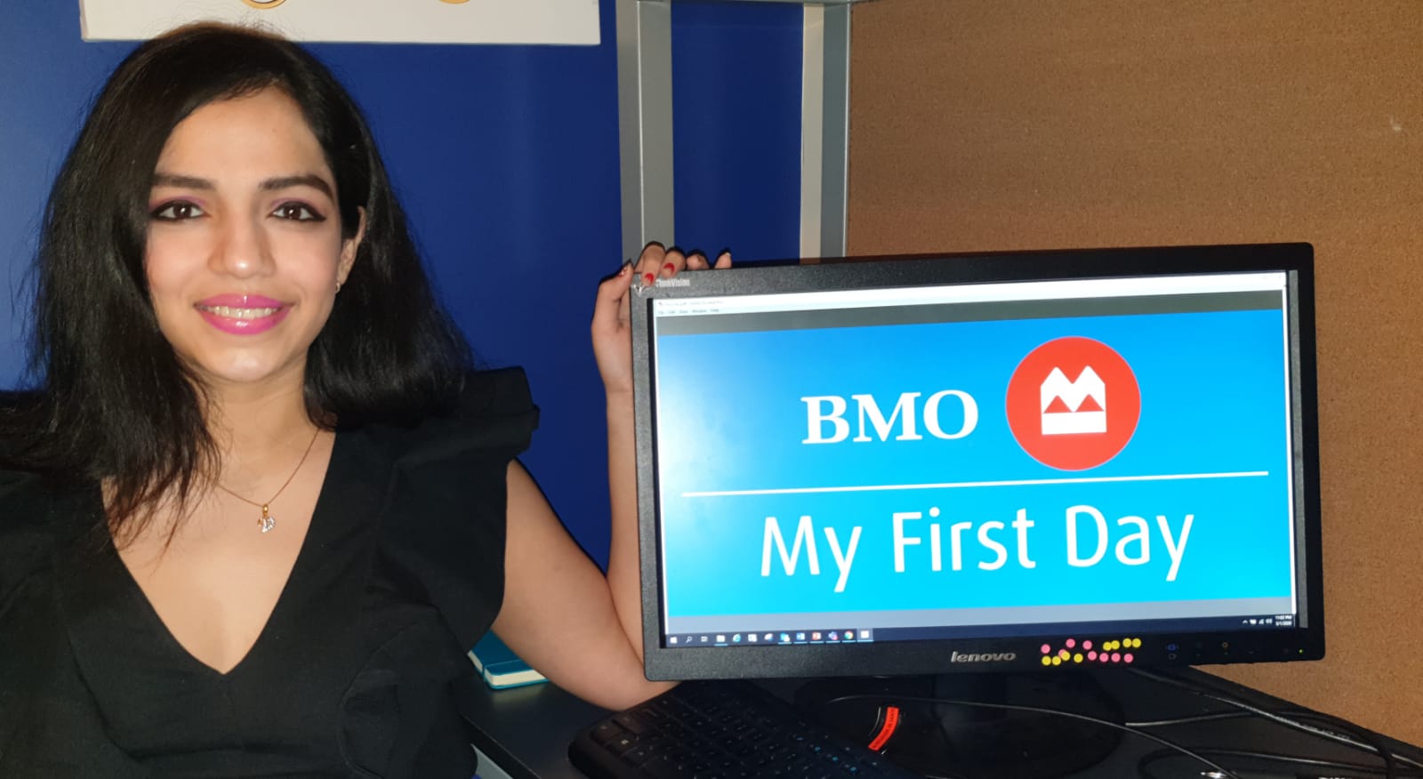 A photo of Vrushti sitting beside a computer showing an image of the BMO logo and the text, &quot;My First Day.&quot;