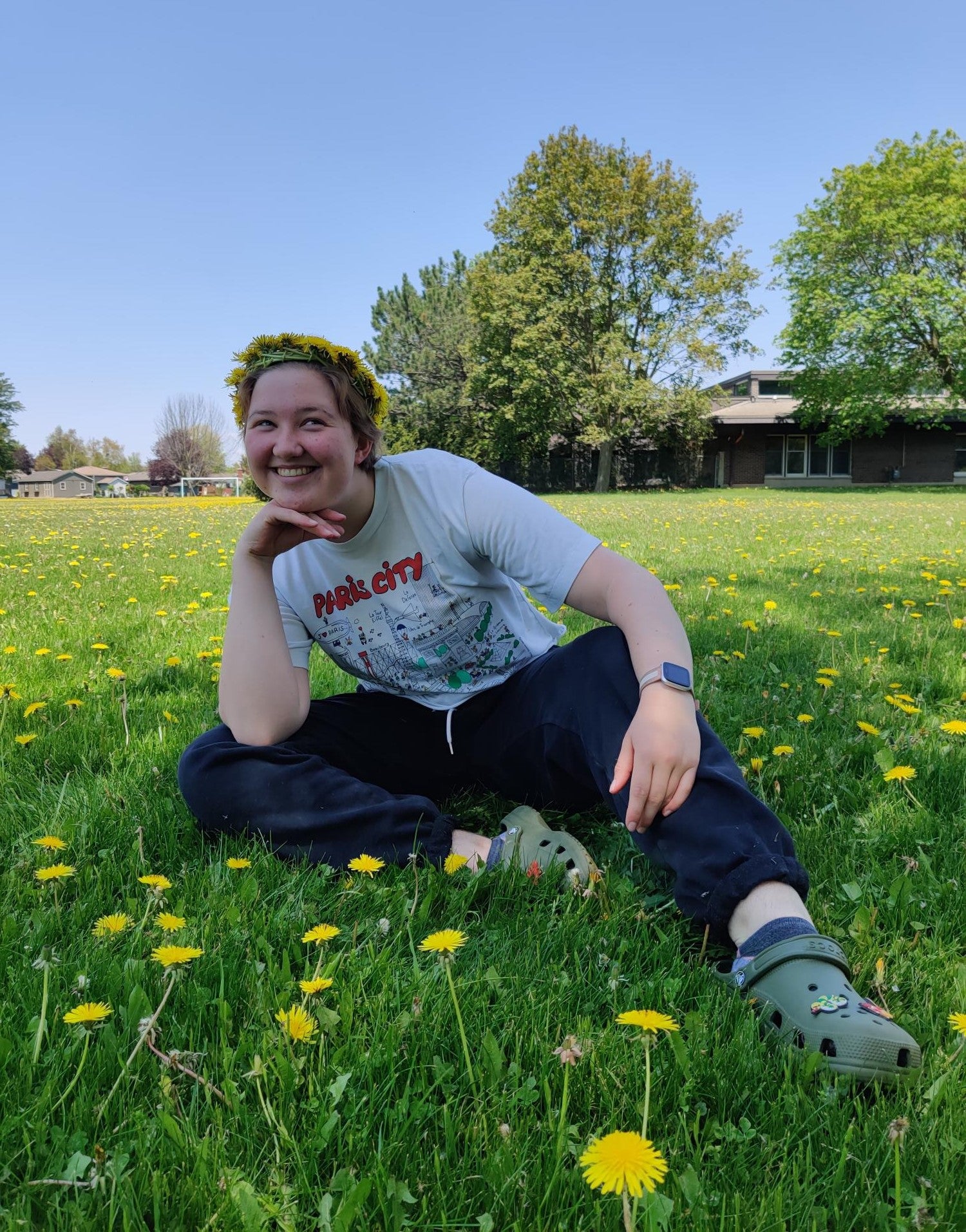 A photo of Charlie sitting outside on the grass and smiling, with their hand underneath their chin.
