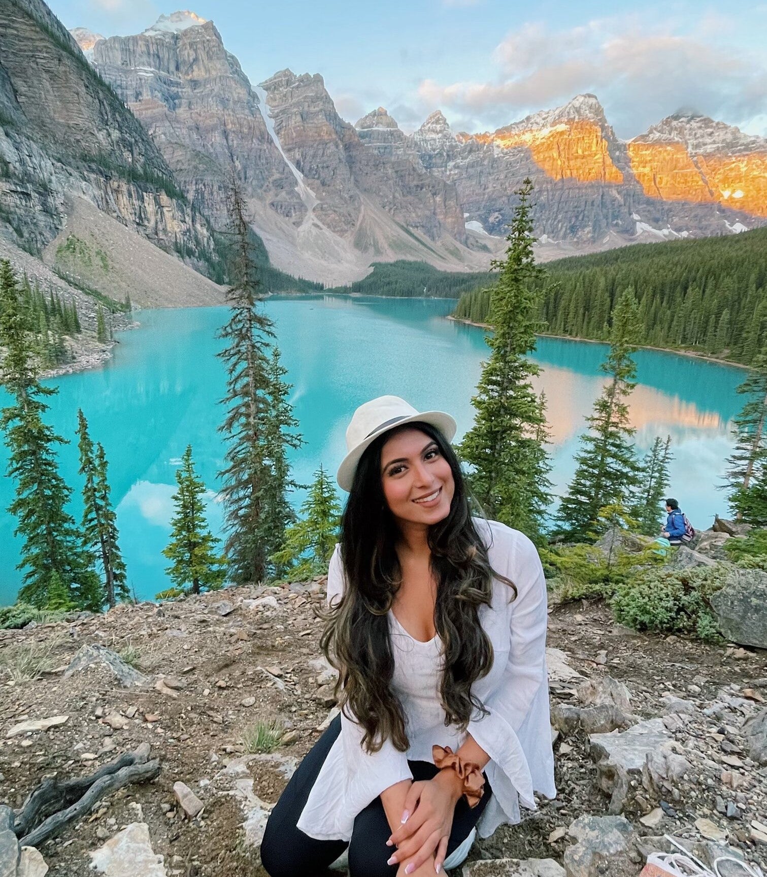 A photo of Rashmi smiling while sitting outside on the ground, with pine trees, water, and mountains in the background.