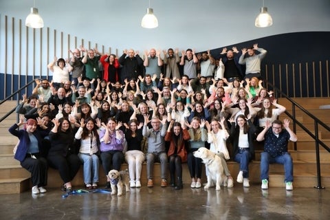 A large group of people pose together for a team photo in an indoor event space, with several participants raising their arms and two dogs sitting at the front.