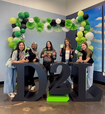 Five people stand behind large "D2L" letters, smiling and holding drinks in front of a green, white and black balloon arch at a D2L office celebration.