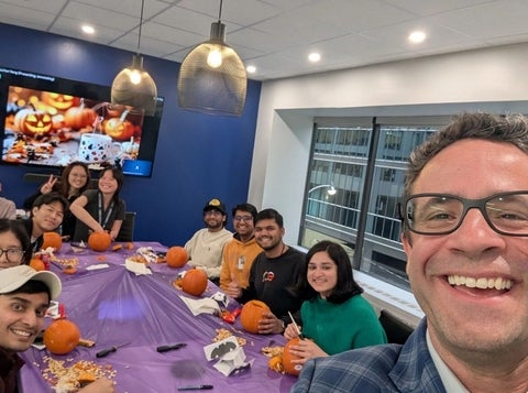 A group of coworkers gathered around a table decorated with pumpkins and Halloween treats, smiling for a selfie during a team celebration.
