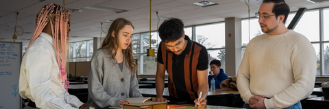 Three students writing with staff standing around a table