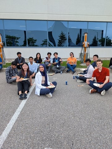 A group of students sitting together outdoors in a parking lot during a team gathering, smiling for a photo in front of an office building.