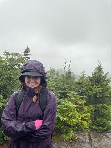 Tara standing on a hiking trail in rainy weather, wearing a waterproof jacket with trees and foggy greenery in the background.