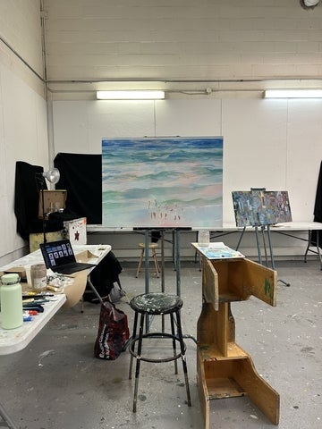 An art studio workspace with paintings on easels, a laptop on a table, and painting supplies arranged around the room.